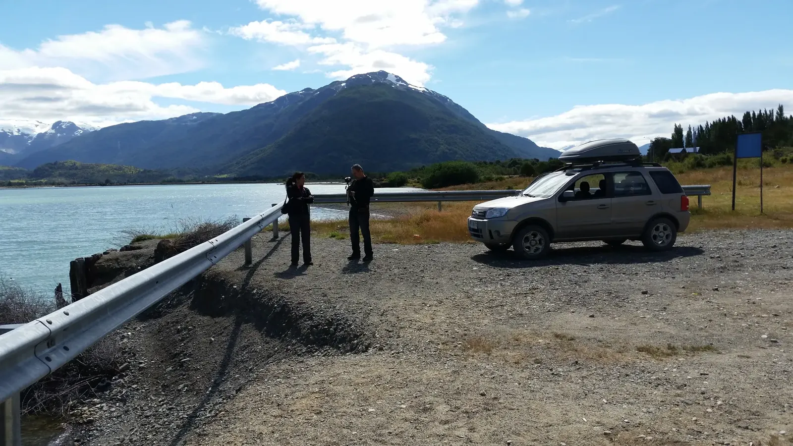 Dos personas junto a un auto detenido frente a un lago y montañas