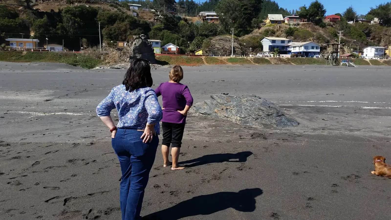 Dos mujeres caminan por una playa de arena oscura frente a un pueblo